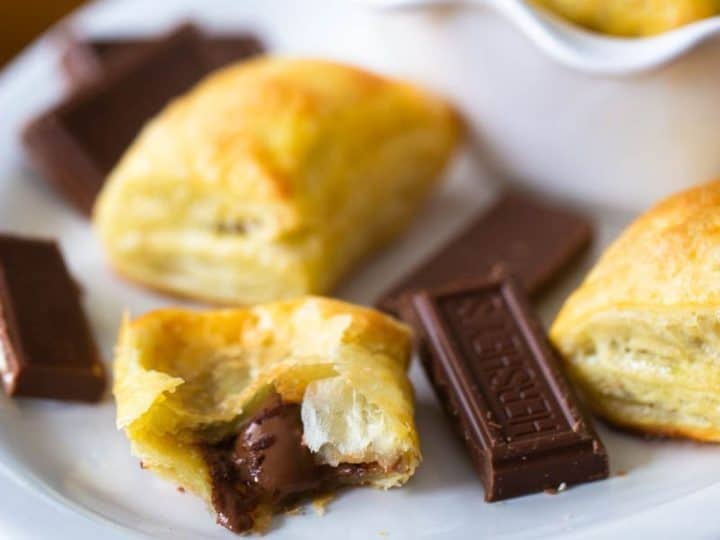 A white platter of homemade chocolate croissants made from puff pastry. One has been broken in half to show the melted chocolate filling, there's a Hershey's square of chocolate next to it.