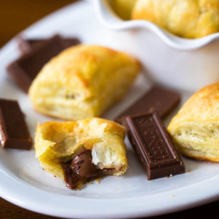 A white platter of homemade chocolate croissants made from puff pastry. One has been broken in half to show the melted chocolate filling, there's a Hershey's square of chocolate next to it.