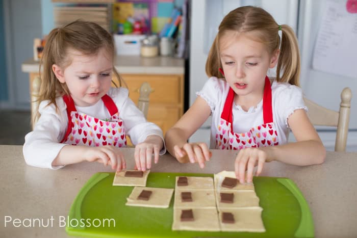 Two little girls wearing Valentine's Day aprons are folding puff pastry dough over chocolate to form chocolate croissants.