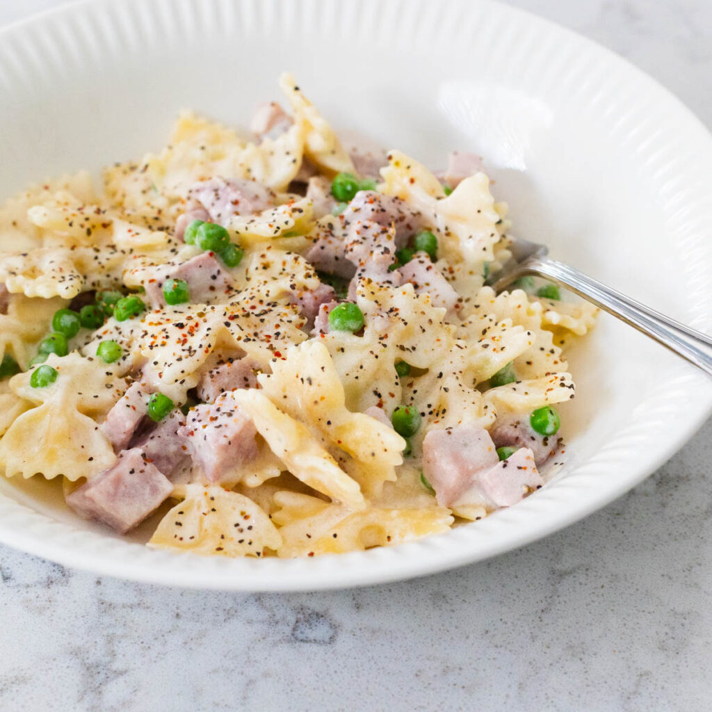 A white bowl filled with farfalle pasta, cubed ham, and peas to show the ham and peas alfredo recipe.