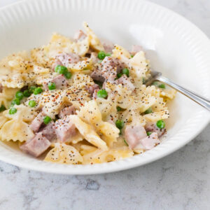 A white bowl filled with farfalle pasta, cubed ham, and peas to show the ham and peas alfredo recipe.