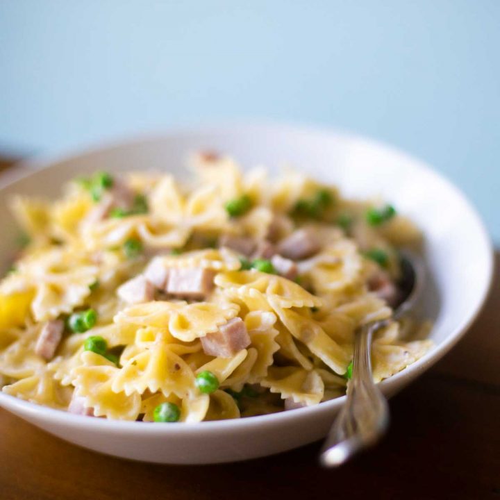 A white bowl filled with farfalle pasta, cubed ham, and peas to show the ham and peas alfredo recipe.