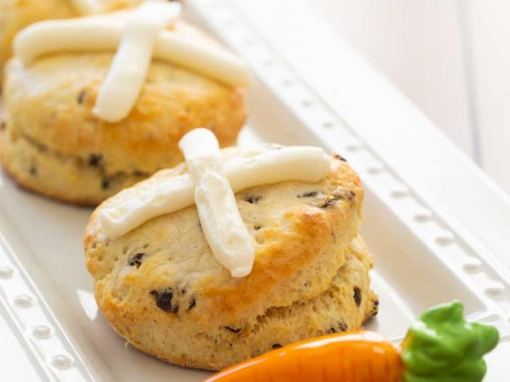 A platter of hot cross biscuits has a ceramic carrot accessory. Each biscuit has a cream cheese frosting cross on the top and currants inside.