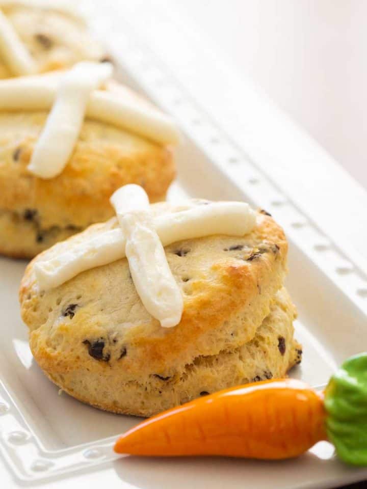 A platter of hot cross biscuits has a ceramic carrot accessory. Each biscuit has a cream cheese frosting cross on the top and currants inside.