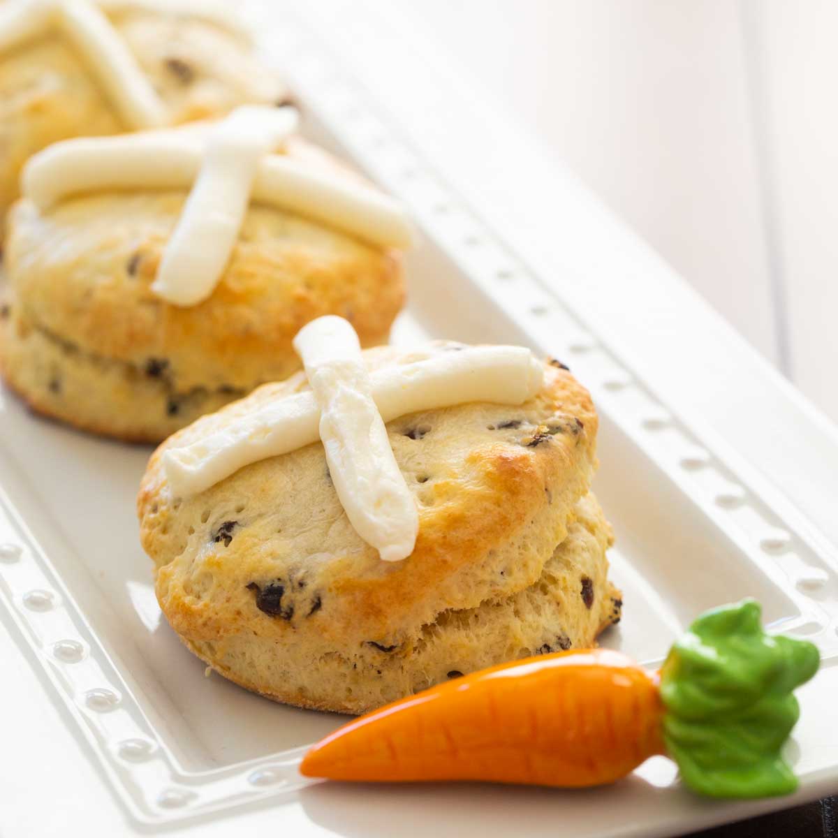 A platter of hot cross biscuits has a ceramic carrot accessory. Each biscuit has a cream cheese frosting cross on the top and currants inside.