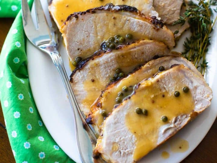 A white platter with slices of roasted pork drizzled with green peppercorn sauce has sprinkles of peppercorns and a bunch of fresh thyme. There's a fork and a green napkin.