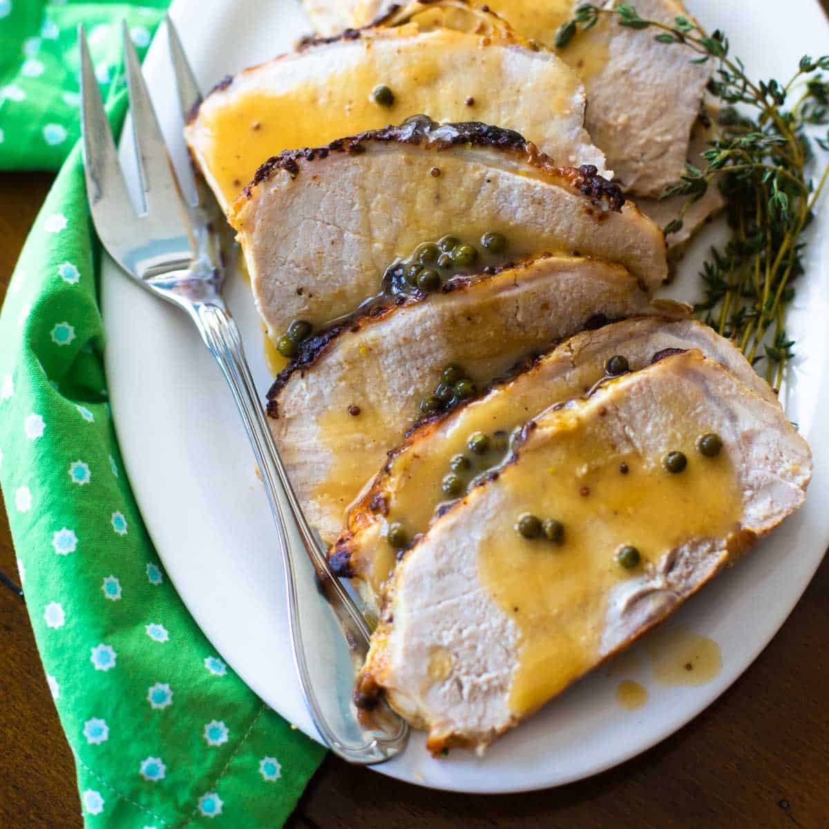 A white platter with slices of roasted pork drizzled with green peppercorn sauce has sprinkles of peppercorns and a bunch of fresh thyme. There's a fork and a green napkin.