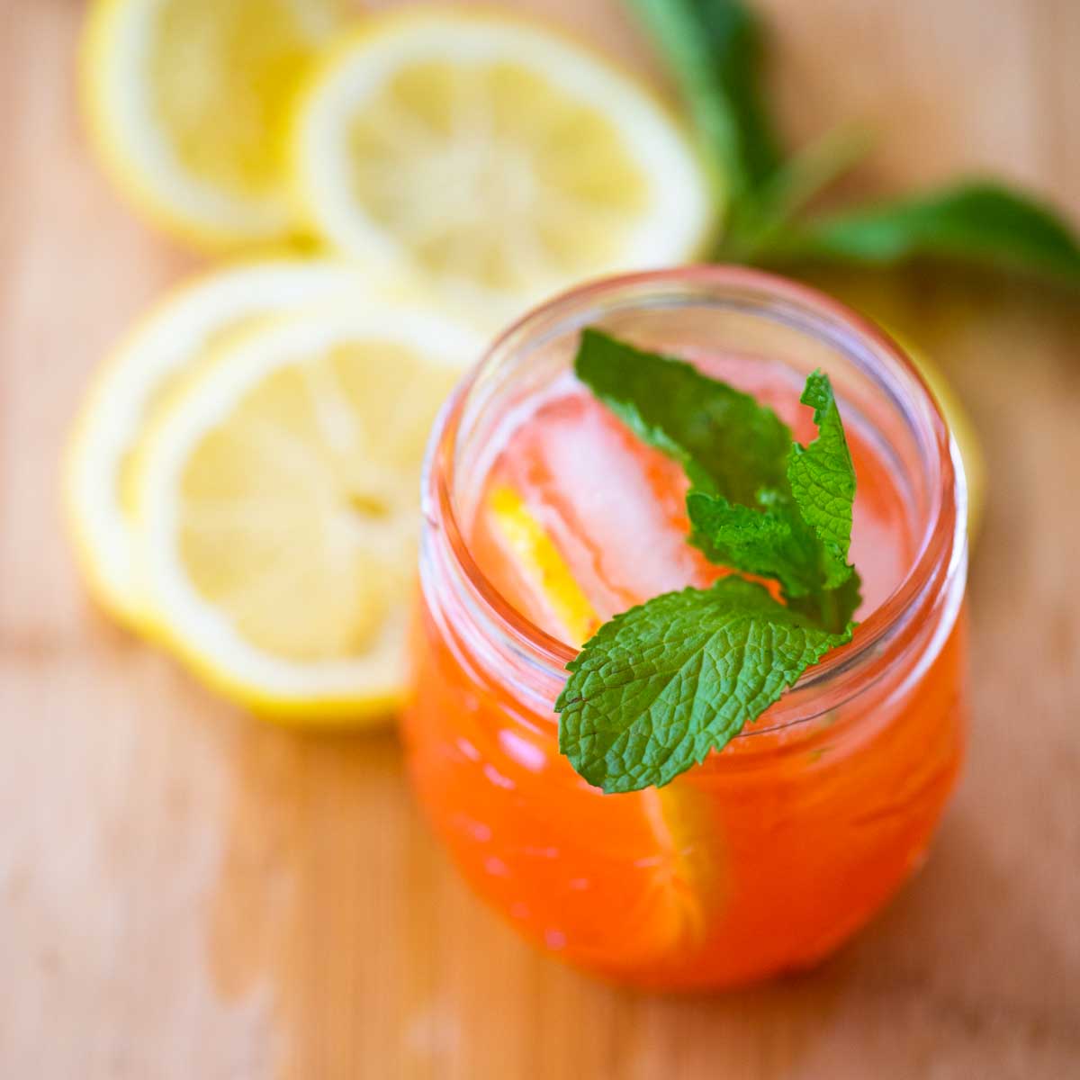 A glass of strawberry lemonade has a fresh sprig of mint on the top for garnish and sits next to several slices of fresh lemon.
