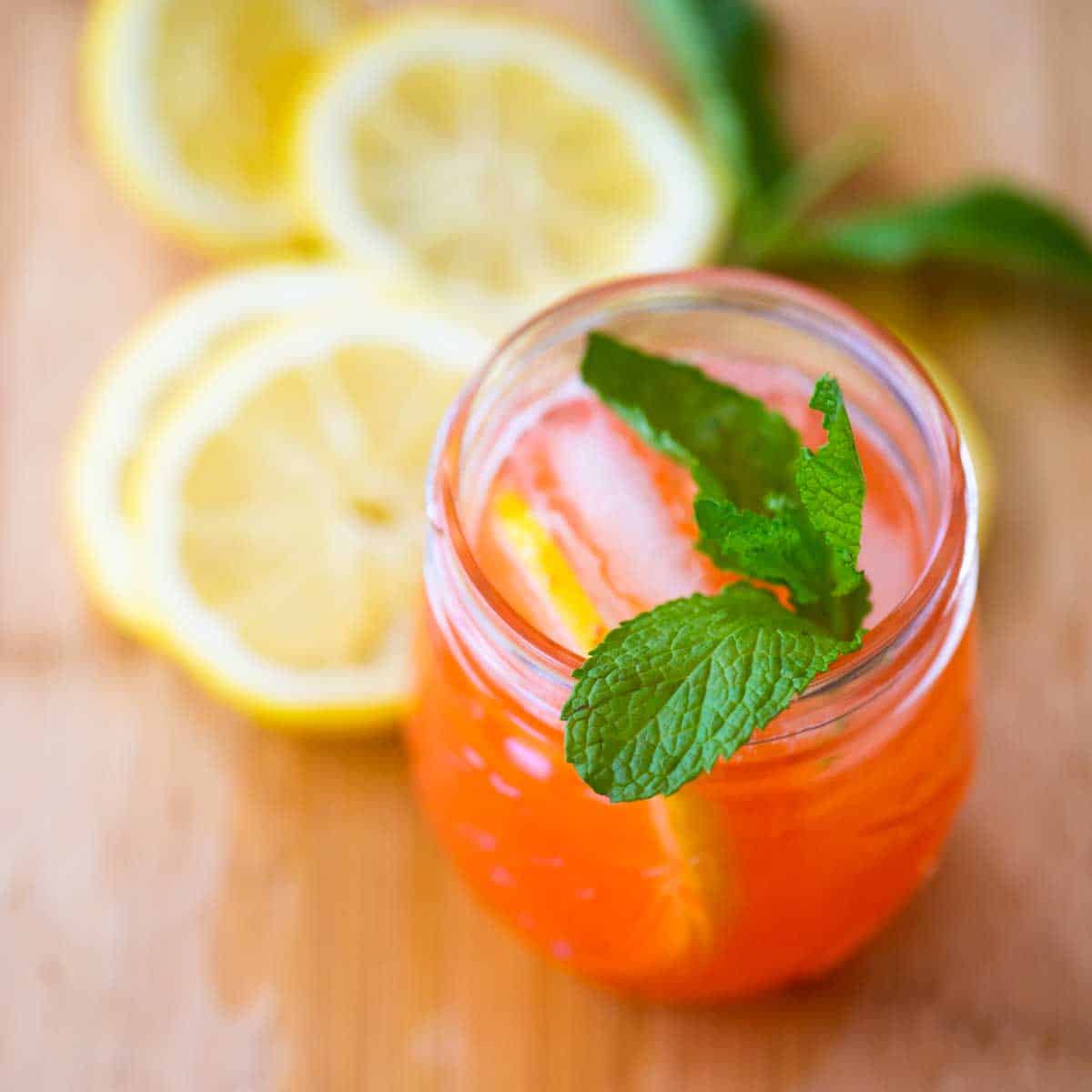 A glass of strawberry lemonade has a fresh sprig of mint on the top for garnish and sits next to several slices of fresh lemon.