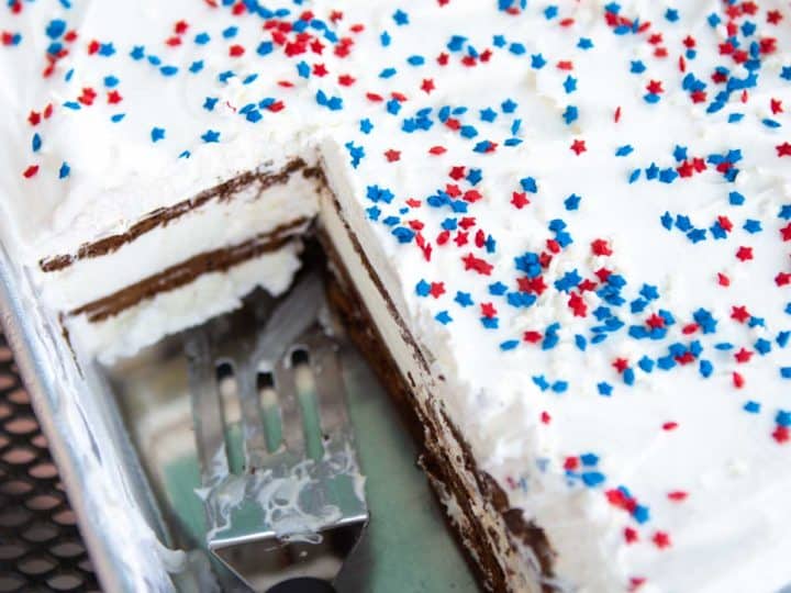 A metal pan has an ice cream cake with red and white sprinkles over the top. A slice is missing so you can see the layers of ice cream sandwiches inside.