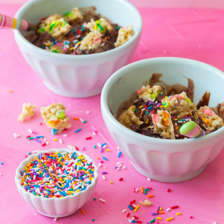 Two bowls of chocolate ice cream with rainbow sprinkles scattered all around. The sundaes are topped with chopped up Lucky Charms cereal bars.