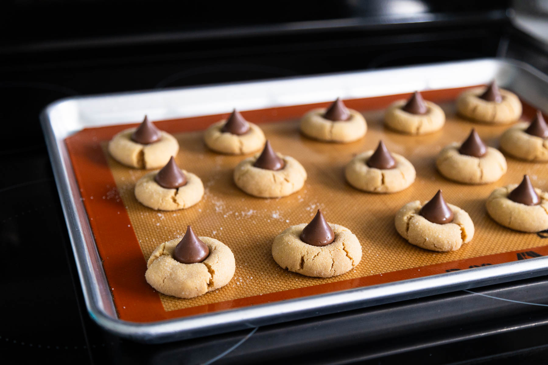 The baking pan of peanut butter blossoms is on the stovetop and the Hershey kisses have just been added to the tops.