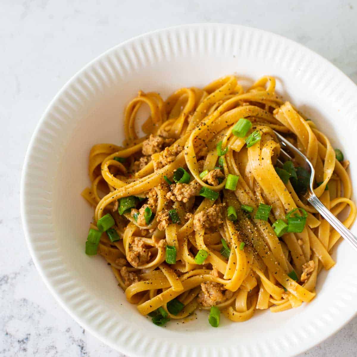 A white bowl has a serving of spicy peanut butter noodles with ground pork and green onions. 