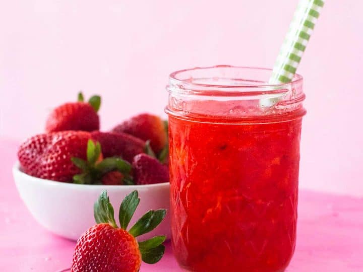 A mason jar filled with homemade strawberry freezer jam has a green plaid spoon sticking out of the top and sits next to a bowl of fresh strawberries.