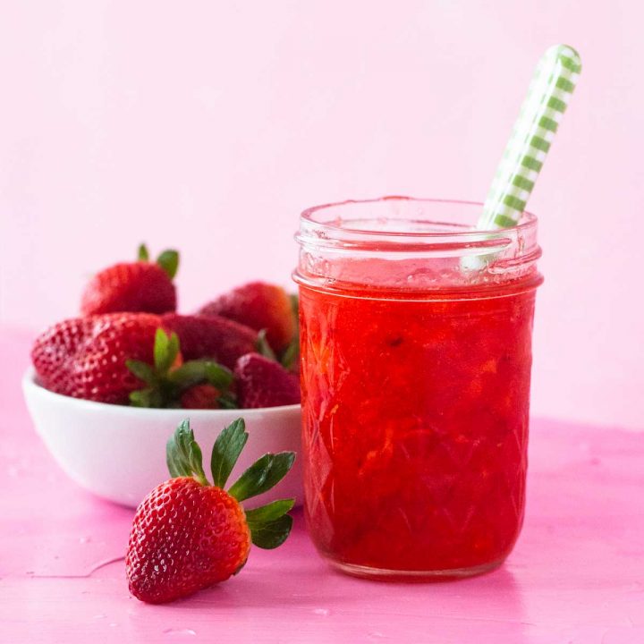 A mason jar filled with homemade strawberry freezer jam has a green plaid spoon sticking out of the top and sits next to a bowl of fresh strawberries.