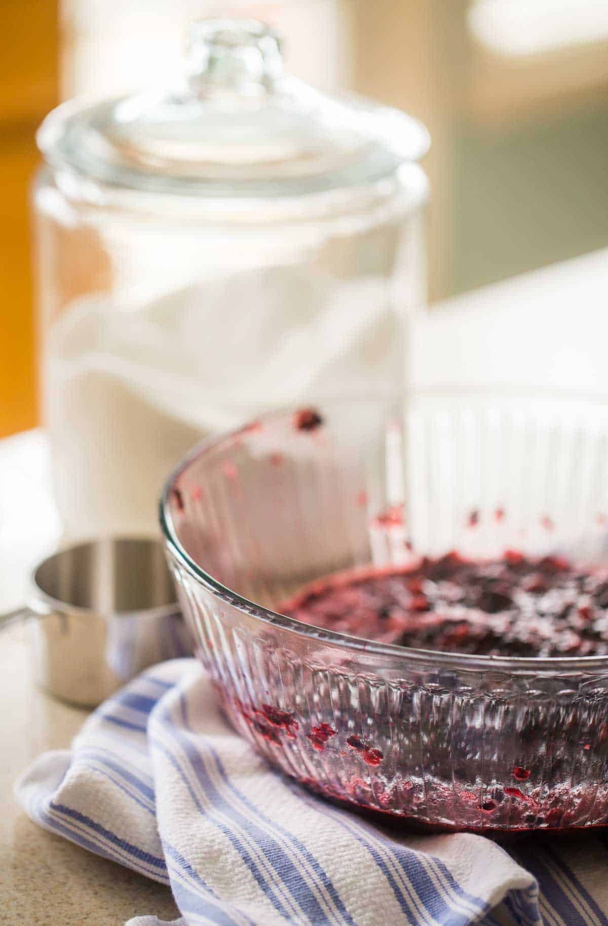 The mixing bowl is filled with crushed blackberries and is sitting next to a container of sugar.