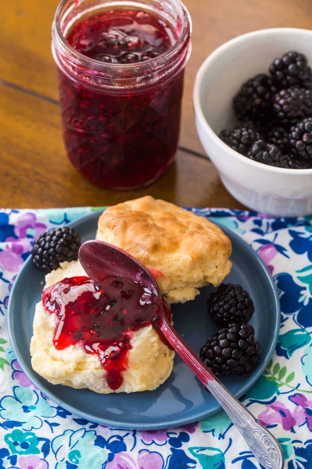 The jam has been spooned onto a fresh baked biscuit. The jelly jar is in the background next to some fresh blackberries.