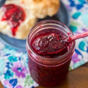 A mason jar with blackberry freezer jam and a spoon. A split biscuit topped with jam sits in the back.