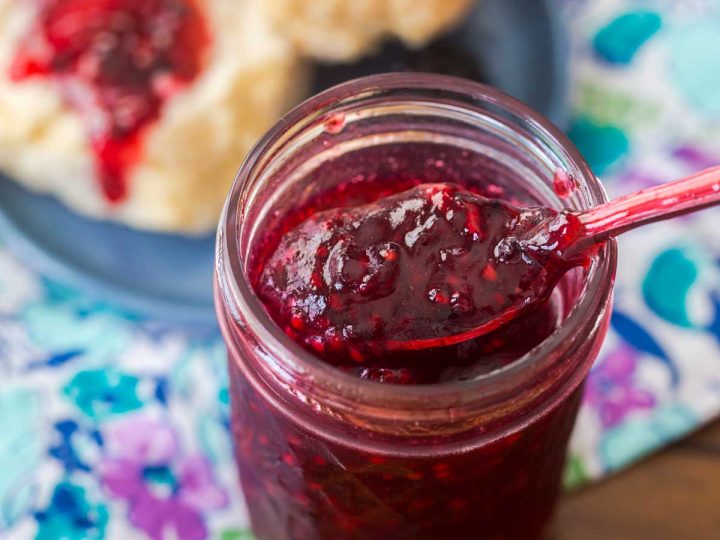 A mason jar with blackberry freezer jam and a spoon. A split biscuit topped with jam sits in the back.