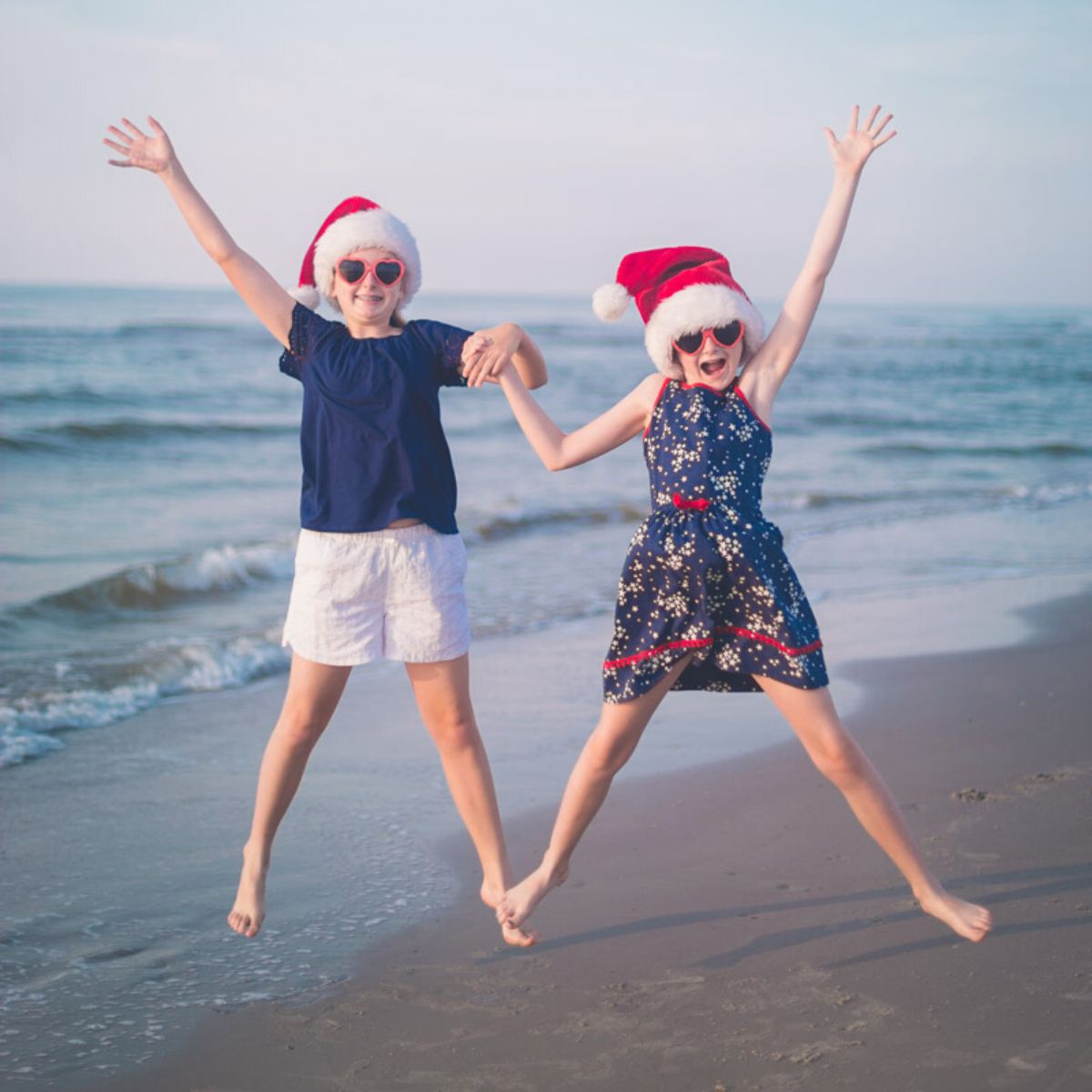 Two young girls wearing Santa hats jumping on a beach