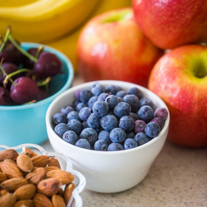A bowl of fresh blueberries sits next to a bowl of cherries, a pile of red apples, fresh bananas, and a bowl of whole almonds.