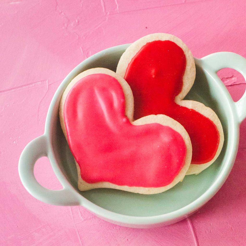 Two heart shaped cut out sugar cookies decorated with pink and red icing sit in an aqua dish on a pink background.