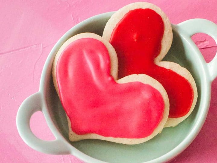 Two heart shaped cut out sugar cookies decorated with pink and red icing sit in an aqua dish on a pink background.