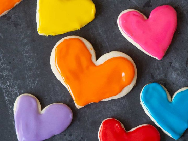 Heart-shaped frosted sugar cookies in rainbow colors against a black chalkboard.