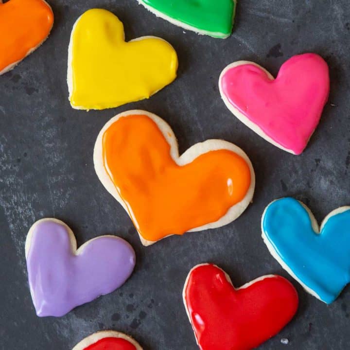Heart-shaped frosted sugar cookies in rainbow colors against a black chalkboard.