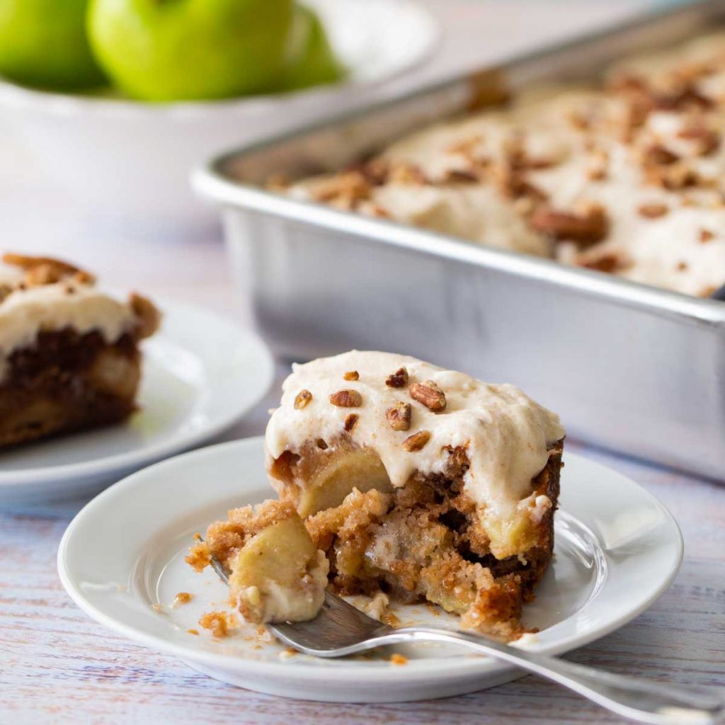 A square slice of apple cake topped with soft frosting and pecans sits in front of the metal baking pan it came from. A bite is missing.