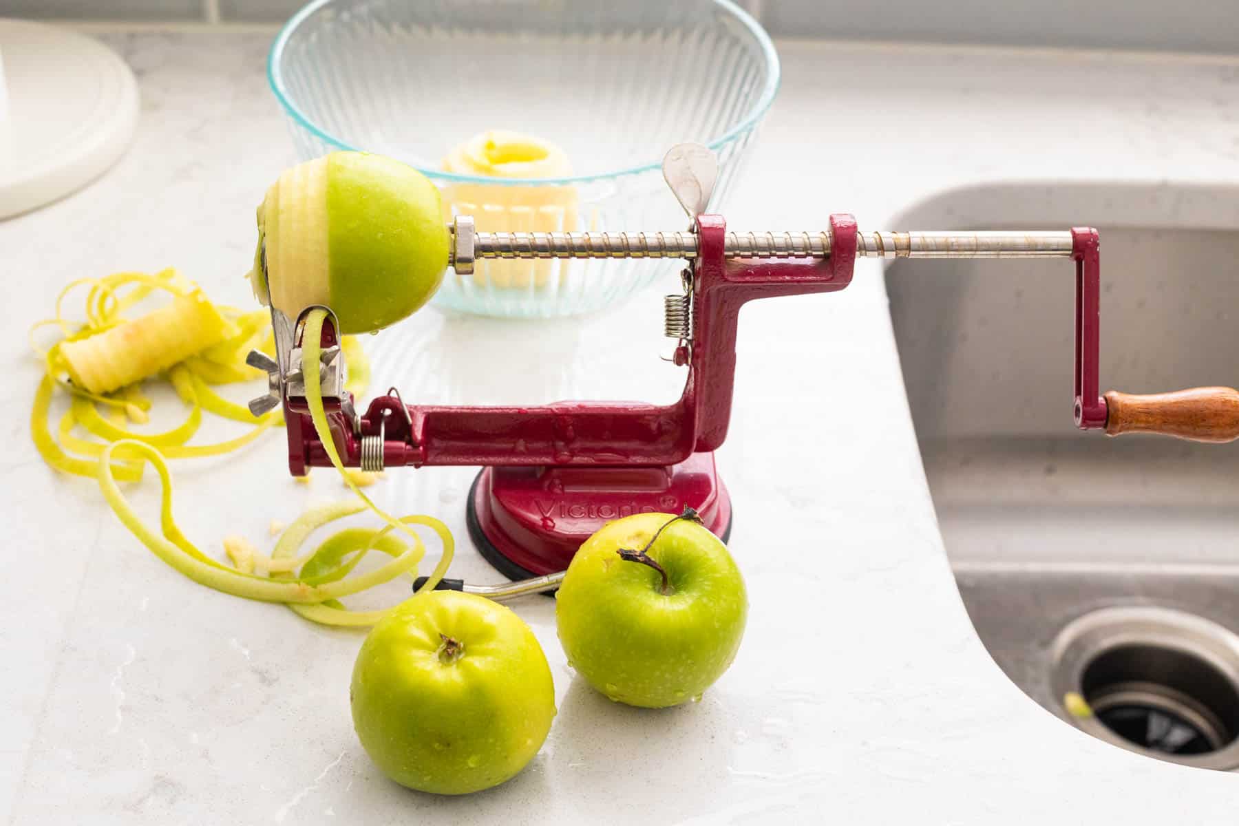 The apple peeler tool is arranged by the kitchen sink and the green apples are in the process of being spiral-peeled.