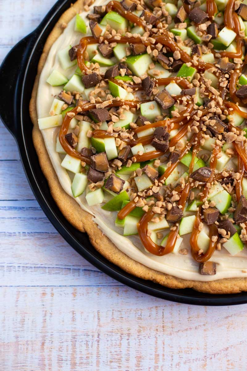 The sugar cookie crust is topped with cream cheese frosting, chopped green apples, and chopped candies and caramel.