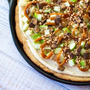 A sugar cookie baked in a pie skillet is topped with fresh chopped apples, chopped snickers candies, and is drizzled with caramel sauce over the top.