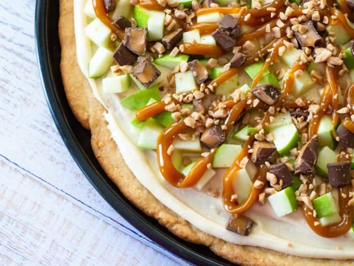A sugar cookie baked in a pie skillet is topped with fresh chopped apples, chopped snickers candies, and is drizzled with caramel sauce over the top.
