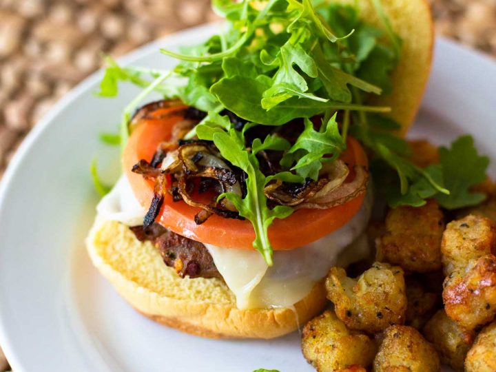 An oven baked turkey burger sits on an open faced bun and is topped with fresh tomato slices, green arugula leaves, and a melted slice of cheese. Tater tots sit next to it for a side dish.