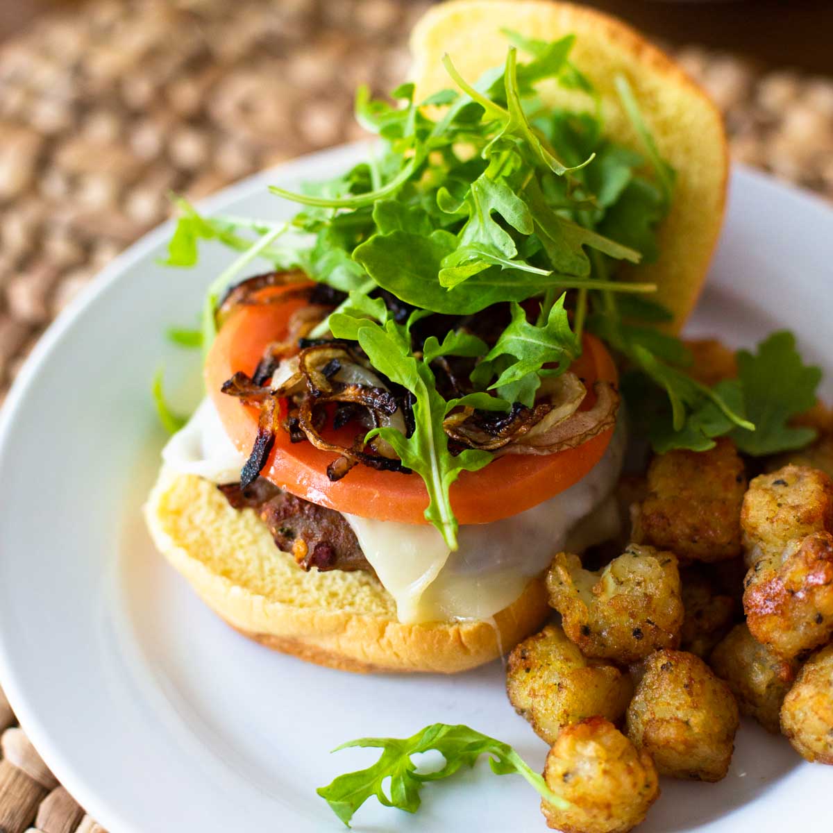 An oven baked turkey burger sits on an open faced bun and is topped with fresh tomato slices, green arugula leaves, and a melted slice of cheese. Tater tots sit next to it for a side dish.