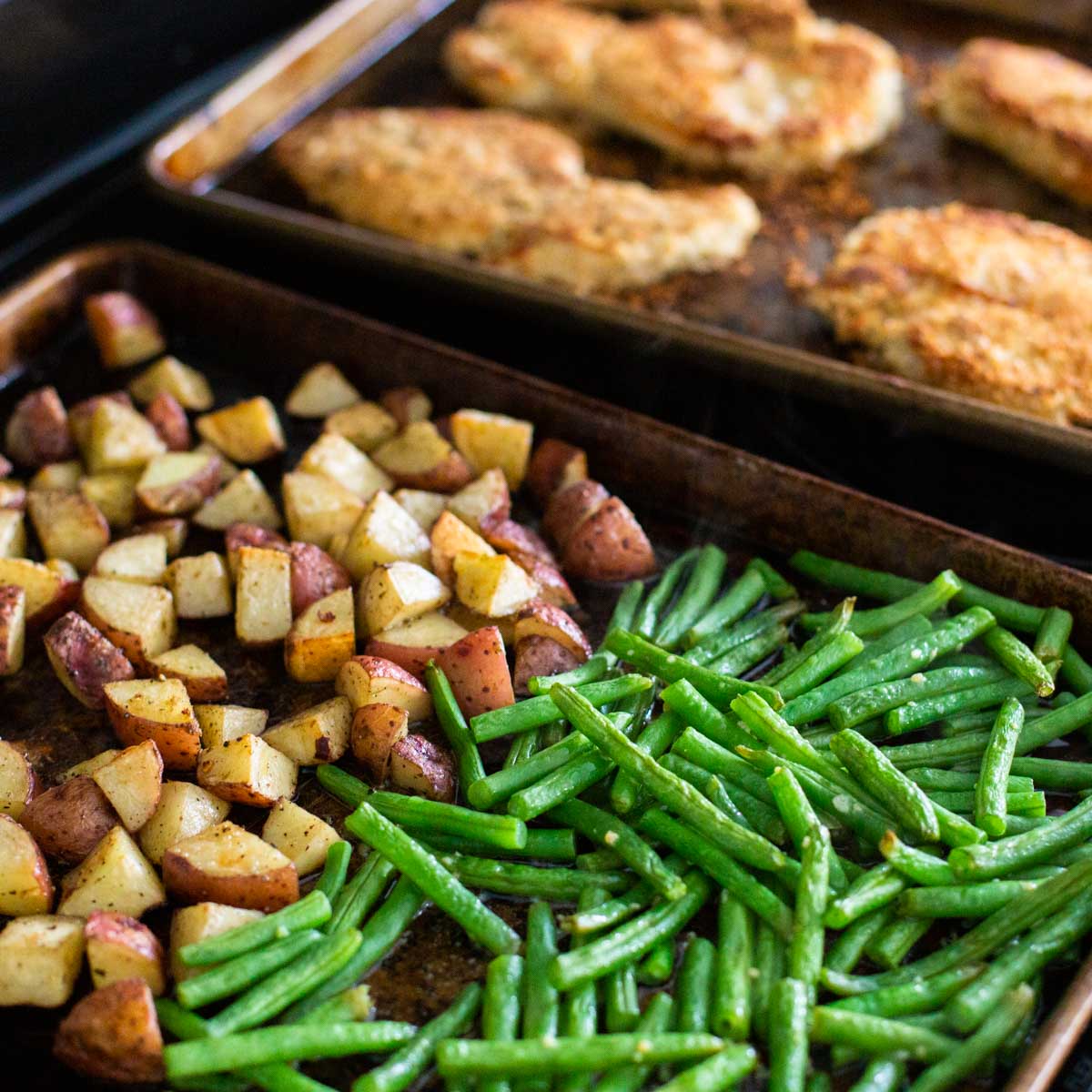Two sheet pans hold roasted green beans, roasted potatoes, and a breaded sheet pan chicken cutlet.