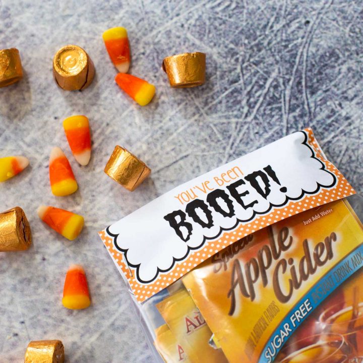 The You've Been Booed printable is stapled to a packet of apple cider with candy corn and Rolos scattered in the background.