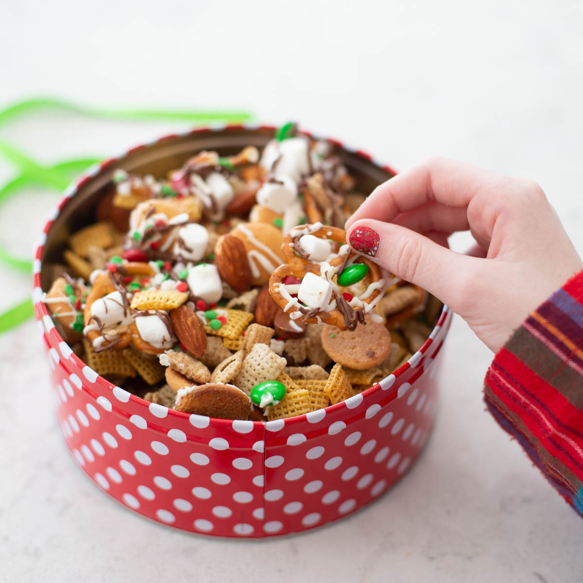 A young girl in Christmas pajamas reaches for a bite of the Christmas chex mix in a red serving container.