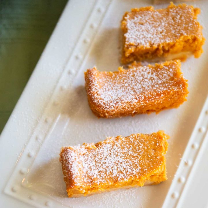 Slices of homemade pumpkin gooey butter cakes are dusted with powdered sugar on a white platter.