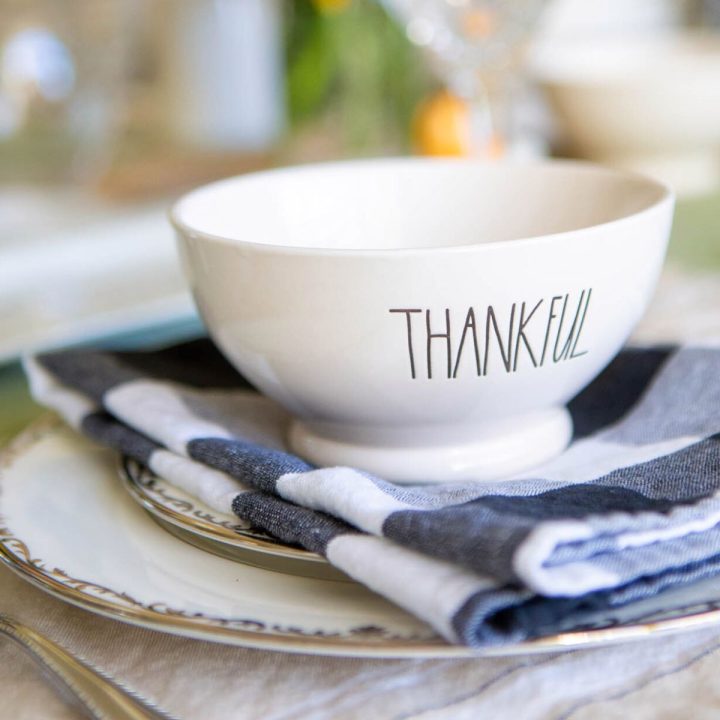 A china plate has a black and white napkin placed on top and a small white bowl that says "Thankful" sits on top.