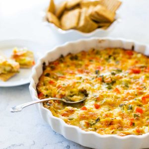 A dish of baked jalapeño artichoke dip shows diced red peppers and green onions. There's a serving spoon taking a scoop. A bowl of crackers sits in the back.