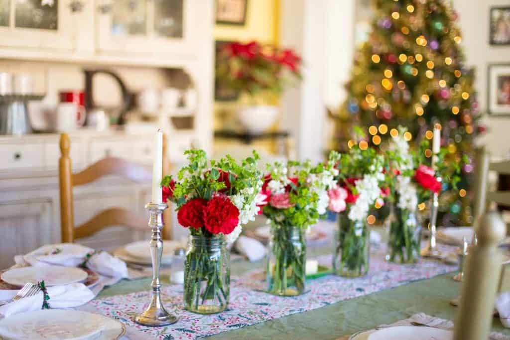 A table is set for Christmas dinner with red and white flowers down the center of the table and a Christmas tree in the background.