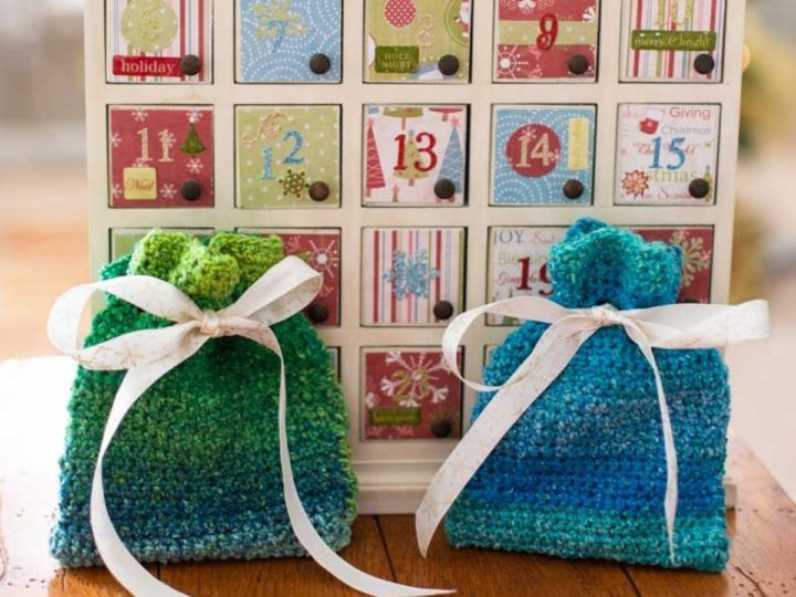 A blue and a green crochet gift bag tied close with ribbon sit on a table in front of an Advent calendar.