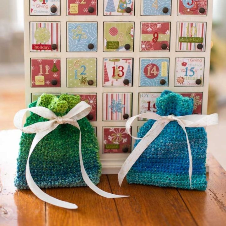 A blue and a green crochet gift bag tied close with ribbon sit on a table in front of an Advent calendar.