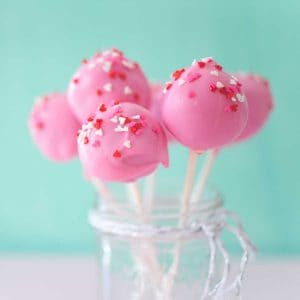 A mason jar filled with pink brownie pops on white sticks with red and white sprinkles.