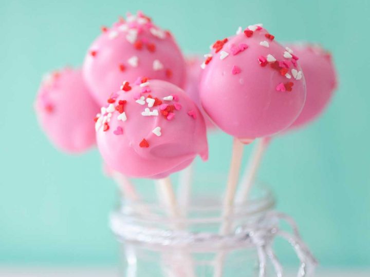 A mason jar filled with pink brownie pops on white sticks with red and white sprinkles.