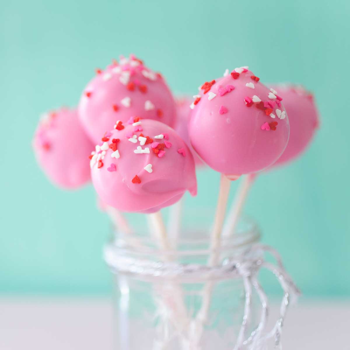 A mason jar filled with pink brownie pops on white sticks with red and white sprinkles.