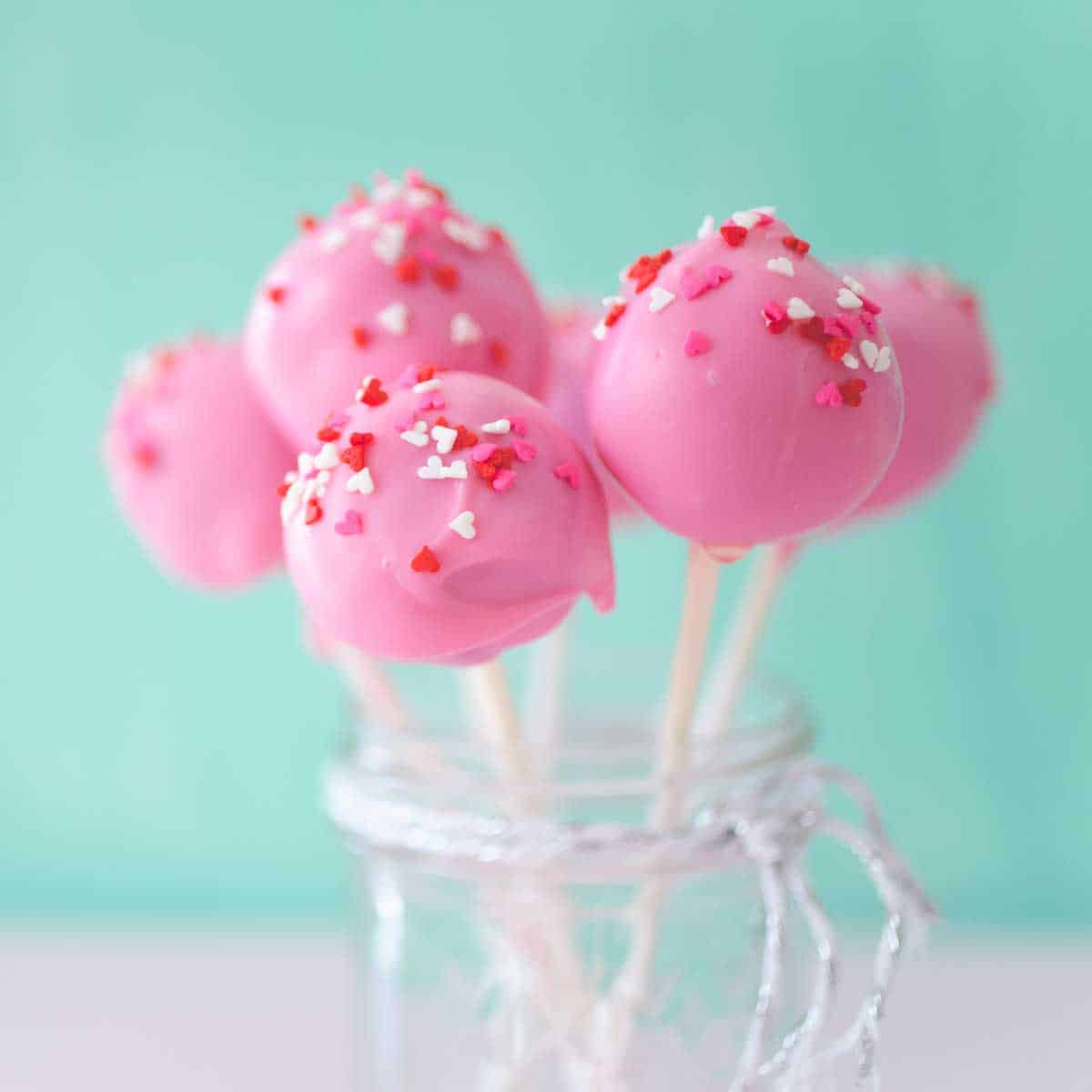 A mason jar filled with pink brownie pops on white sticks with red and white sprinkles.