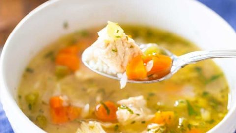 A bowl of homemade chicken soup with chicken and carrots has a spoon scooping up a bite.